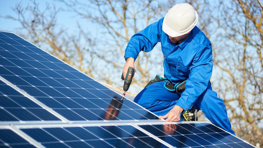 Worker in blue overalls and a white helmet installing solar panelsin Langebaan, with a drill on a rooftop.